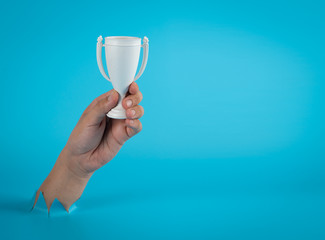 Hand holding white trophy cup through the torn blue plastic background,Winner concept