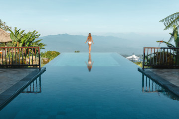 Slim young woman in swimsuit relaxing on edge tropical infinity pool in mountains. Palms around and crystal clean water. Luxury resort on Bali island - obrazy, fototapety, plakaty