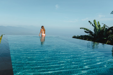 Slim young woman in swimsuit relaxing on edge tropical infinity pool in mountains. Palms around and...