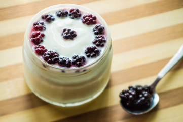 Yogurt in transparent jars with fresh blackberry and strawberry voyen with a spoon, on wooden background