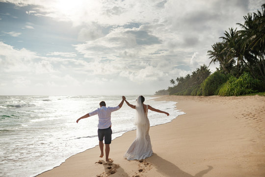Happy bride and groom running on a beautiful tropical sand beach. Couple at beautiful beach wedding - Powered by Adobe