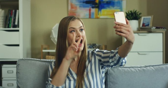 Close up of the young caucasian pretty girl in the striped blouse sitting on the sofa in the cozy room and taking selfie photo on the smartphone. At home.