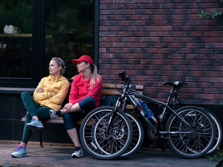 Two young girls in sportswear are resting on a bench in a city park near the entrance to the cafe. Bicycles stand nearby.