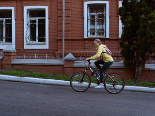 Fototapeta na wymiar A young girl in a yellow jacket with a backpack rides a bicycle on an asphalt road past brick houses.