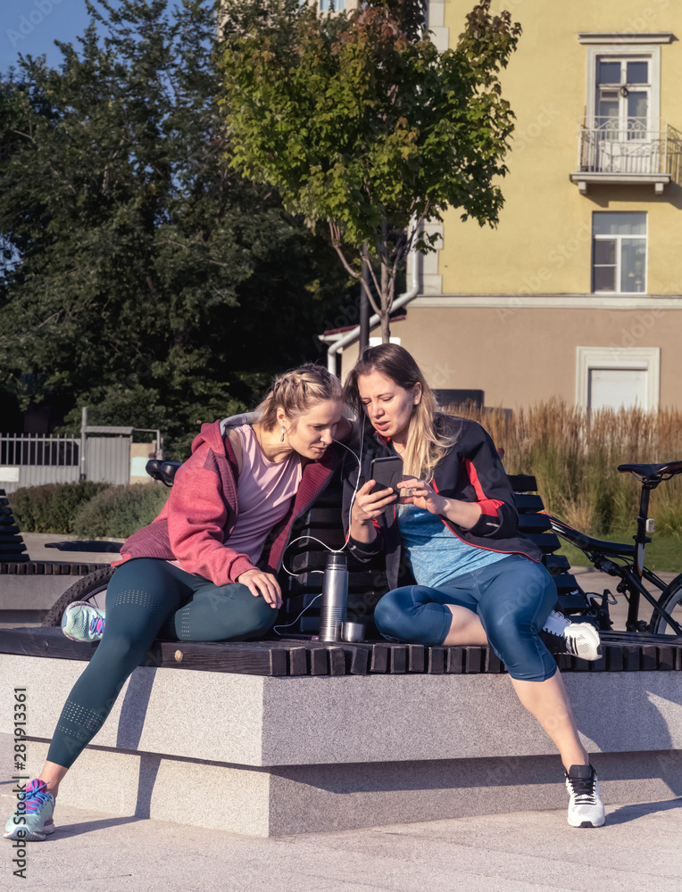 Wall mural two young girls in sportswear are sitting on a bench in a city square and listening to music. they a