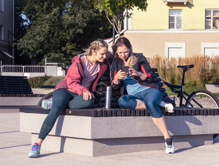 Two young girls in sportswear are sitting on a bench in a city square and listening to music. They are holding phones in their hands. Girls rest after a bike ride.