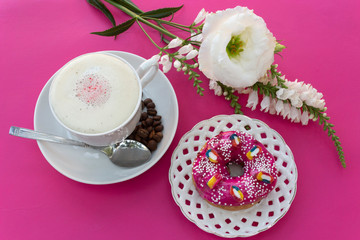A cup of cappuccino coffee with milk froth, a bright pink strawberry donut on a white openwork plate on a pink-purple table where there are white flowers - this is a bright breakfast or coffee break.