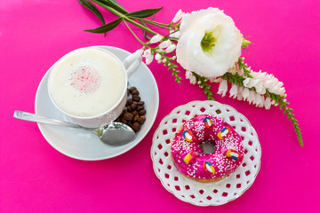 A cup of cappuccino coffee with milk froth, a bright pink strawberry donut on a white openwork plate on a pink-purple table where there are white flowers - this is a bright breakfast or coffee break.