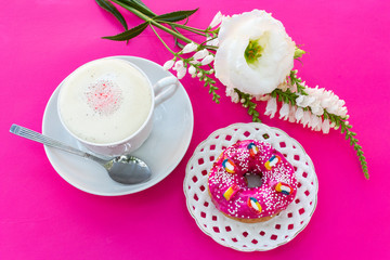 A cup of cappuccino coffee with milk froth, a bright pink strawberry donut on a white openwork plate on a pink-purple table where there are white flowers - this is a bright breakfast or coffee break.