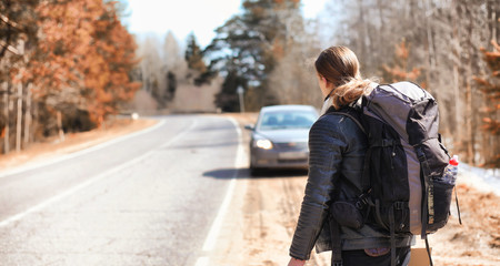 A young man is hitchhiking around the country. The man is trying to catch a passing car for traveling. The man with the backpack went hitchhiking to the south.