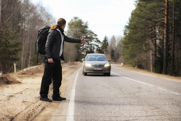 A young man is hitchhiking around the country. The man is trying to catch a passing car for traveling. The man with the backpack went hitchhiking to the south.