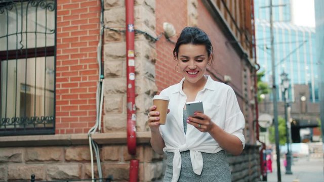 Portrait of attractive girl using smartphone holding take out coffee walking outdoors smiling in the city street. People, gadgets and moden lifestyle concept.