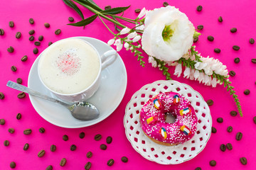 A cup of cappuccino coffee with milk foam, a bright pink strawberry donut on a white openwork plate on a purple table, where there are white flowers and coffee beans - this is a bright breakfast 