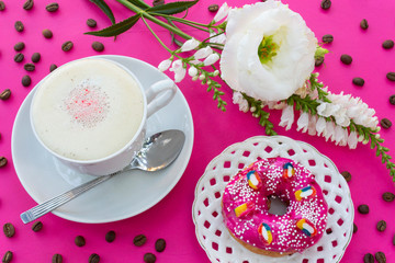 A cup of cappuccino coffee with milk foam, a bright pink strawberry donut on a white openwork plate on a purple table, where there are white flowers and coffee beans - this is a bright breakfast 