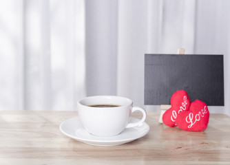 Hot coffee cup with red hearts shape symbol  and black board on wood table  with white curtain background