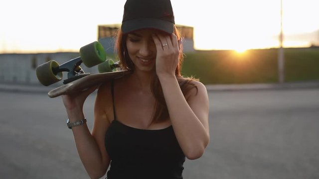 Cool woman skateboarder carries her skateboard on a shoulder at sunset