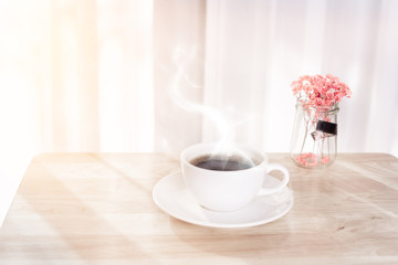White coffee cup with smoke, red dried flower on wooden office desk  with morning sunlight and  soft curtain background