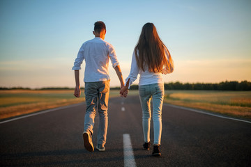 Fototapeta premium Two young people girl and boy holding hands walking along an asphalt road in summer in the countryside. The beginning of the way.