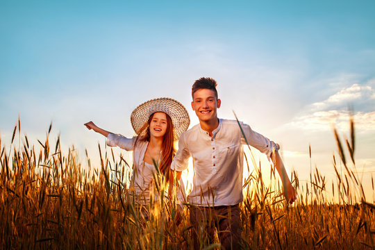 Girl and boy teenagers in white shirts running on a wheat field on a summer evening at sunset. Enjoying nature and freedom. First love, beautiful young couple. Happy children, brother and sister.