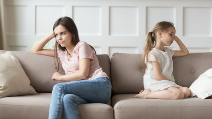 Dissatisfied mother and little daughter sitting on couch looking aside