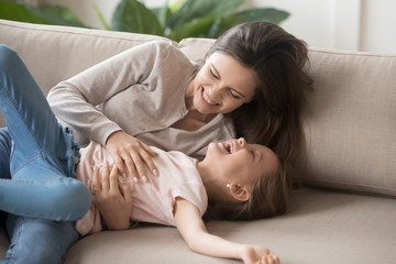 Laughing mother tickling daughter lying on couch in living room