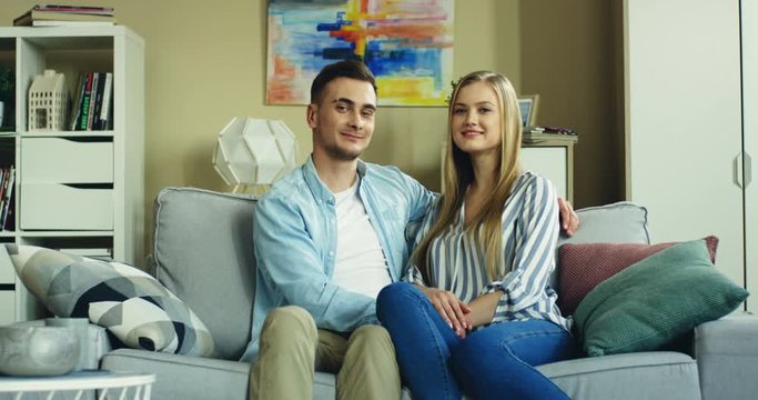 Portrait shot of the romantic happy couple sitting on the couch, looking at each other with love and then smiling to the camera in the nice living room.
