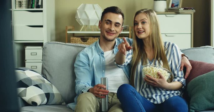 Cheerful Caucasian young couple laughing and talking while watching TV together on the couch athome with a bowl of popcorn.