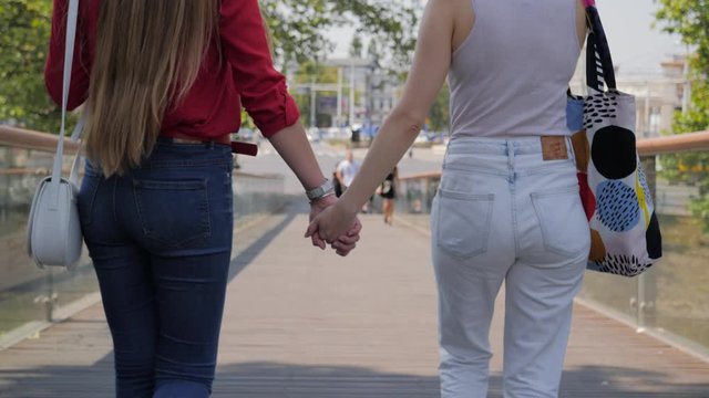 Two young women in love walk together in city holding hands back view