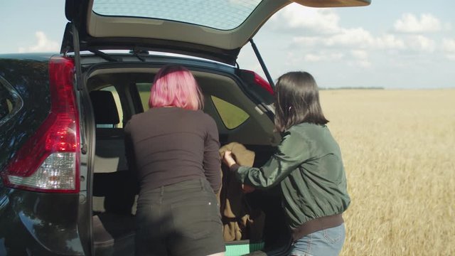 Multiethnic female friends packing for road trip, emotionally discussing needfulness of packed things into luggage near car trunk, throwing away stuffs while preparing for summer vacations travel.