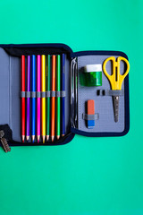Pencils, scissors, an eraser in a school pencil case on a green paper background with copy space.