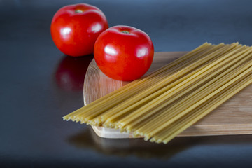 Uncooked whole wheat spaghetti pasta with tomatoes on a cutting board on a dark background.
