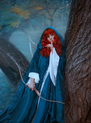 cute red-haired girl in blue cloak standing by tree and looking directly at camera, lady holds...