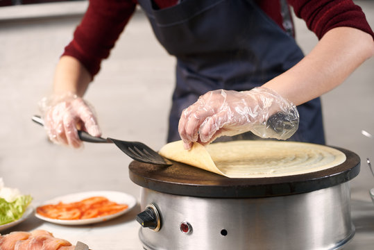 Cropped Woman Chef Hands In Gloves Turn Over Big Pancake With Spatula On Electric Round Cooktop While Baking Process. Lettuce, Salmon And Tomato Slices In Plate On Blurred Light Background. Front View