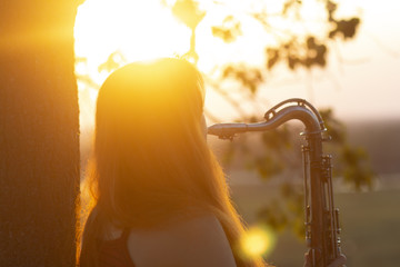silhouette of a red-haired girl standing near a tree and playing a wind instrument at sunset, young...