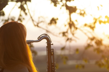 silhouette of a red-haired girl standing near a tree and playing a wind instrument at sunset, young...