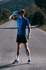 Fit male runner drinking water from a water bottle on an empty road in the woods.