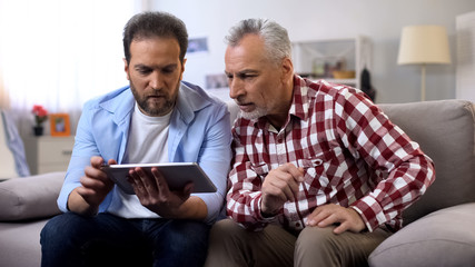 Adult son and senior father watching tutorial videos on tablet, information