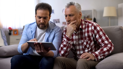 Middle-aged son teaching retiree dad to use banking app on tablet, technologies