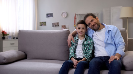 Happy smiling dad and schoolboy son sitting on sofa, looking at camera, family