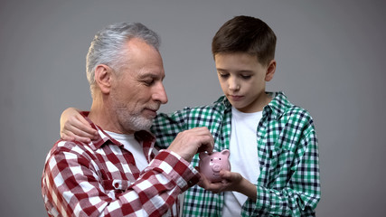 Elderly man putting coin into little boy piggy bank, savings for future, banking