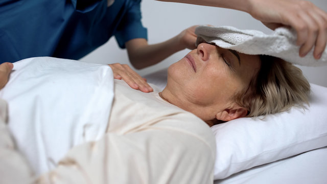 Sick woman suffering from fever and ravings, nurse putting compress on forehead