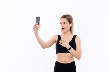 Young sporty blond woman in a black sportswear making selfie with smartphone showing bicep after workout standing over white background. Model listens music during workout with wireless headphones