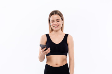 Fototapeta na wymiar Excited young sporty blond woman in a black sportswear with wireless ear buds looking at the screen of a smartphone standing isolated over white background.