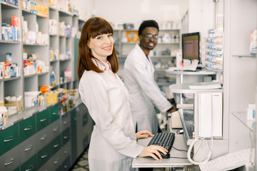 Obraz premium Two young cheerful pharmacists African man and Caucasian woman working together and using computers. Pharmacists searching medicines at pharmacy