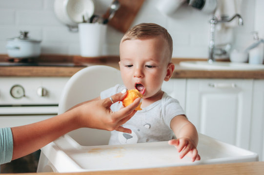 Charming Little Baby Boy Eating First Food Peach At Kitchen. Mom Feeds Child