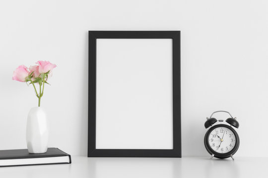 Black frame mockup with workspace accessories  and pink roses in a vase on a white table.Portrait orientation.