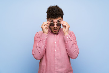 Man with curly hair over isolated blue wall with glasses and surprised