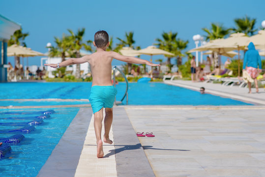 Joyful European boy running along swimming pool at resort in summer. His arms are wide open.