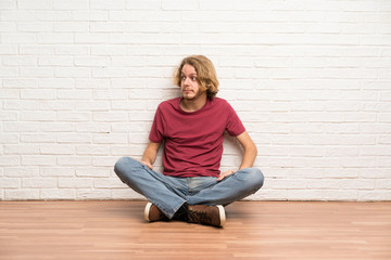 Blonde man sitting on the floor making doubts gesture looking side