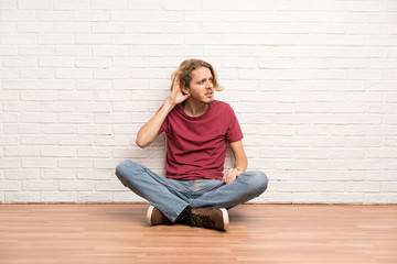 Blonde man sitting on the floor listening to something by putting hand on the ear